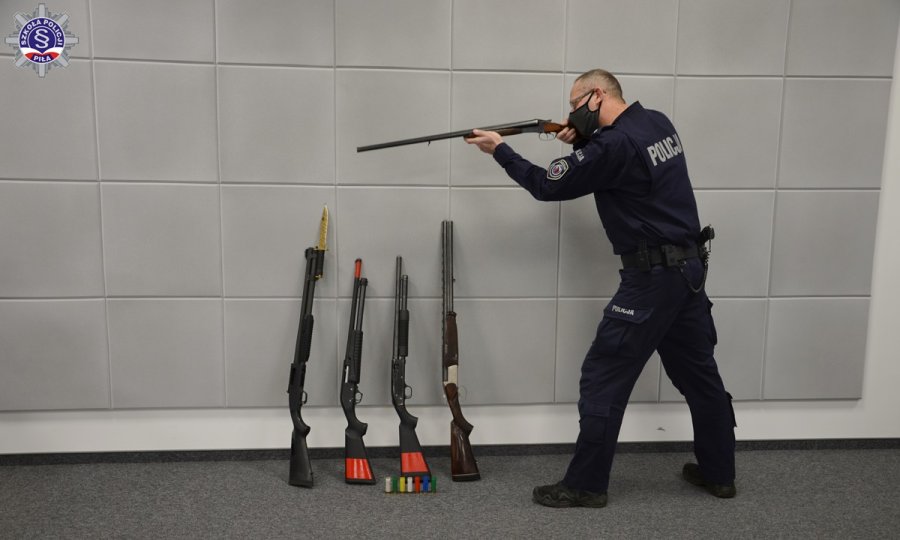 Umundurowany policjant w postawie strzeleckiej z  bronią palną. W tle o ścianę oparte cztery jednostki broni palnej.