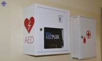 Dwie szafki koloru białego wiszące na ścianie. Pierwsza z napisem AED automatyczny elektryczny defibrylator, druga to podręczna apteczka ze znakiem czerwonego krzyża.