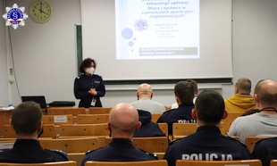 Kobiety i mężczyźni słuchający wykładu umundurowanej policjantki. Na ekranie wyświetlana prezentacja multimedialna.
