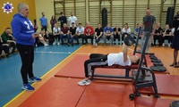 Kobieta w trakcie wykonywania ćwiczenia wyciskania sztangi w pozycji leżącej oraz grupa osób obserwujących zawody.