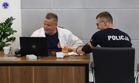 Umundurowany policjant siedzi na krześle przy biurku. Po drugiej stronie biurka mężczyzna w białym fartuchu pisze na klawiaturze komputera.