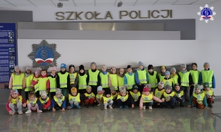 Zdjęcie grupowe przedszkolaków w żółtych kamizelkach przed logo Szkoły Policji w Pile w budynku głównym Szkoły.