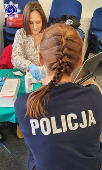Kobieta w policyjnym mundurze, siedząc na fotelu tyłem do obiektywu jest obsługiwana przez pielęgniarkę przy biurku.
