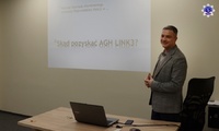 Jeden z prelegentów stojący przy biurku, w tle prezentacja ze slajdem „Skąd pozyskać AGH LINK3?”